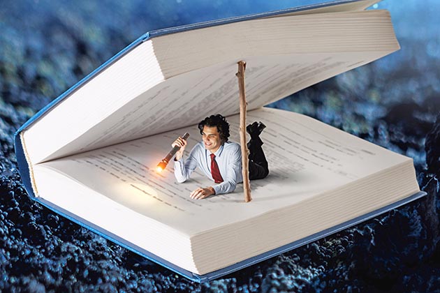 Image of a man holding a flashlight laying inside a gigantic book looking for errors.